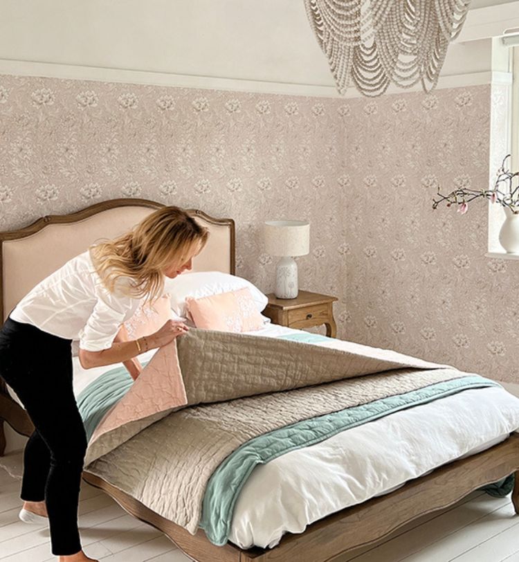 Woman making a bed in a stylish bedroom with floral wallpaper, beige bedding, and a decorative chandelier.