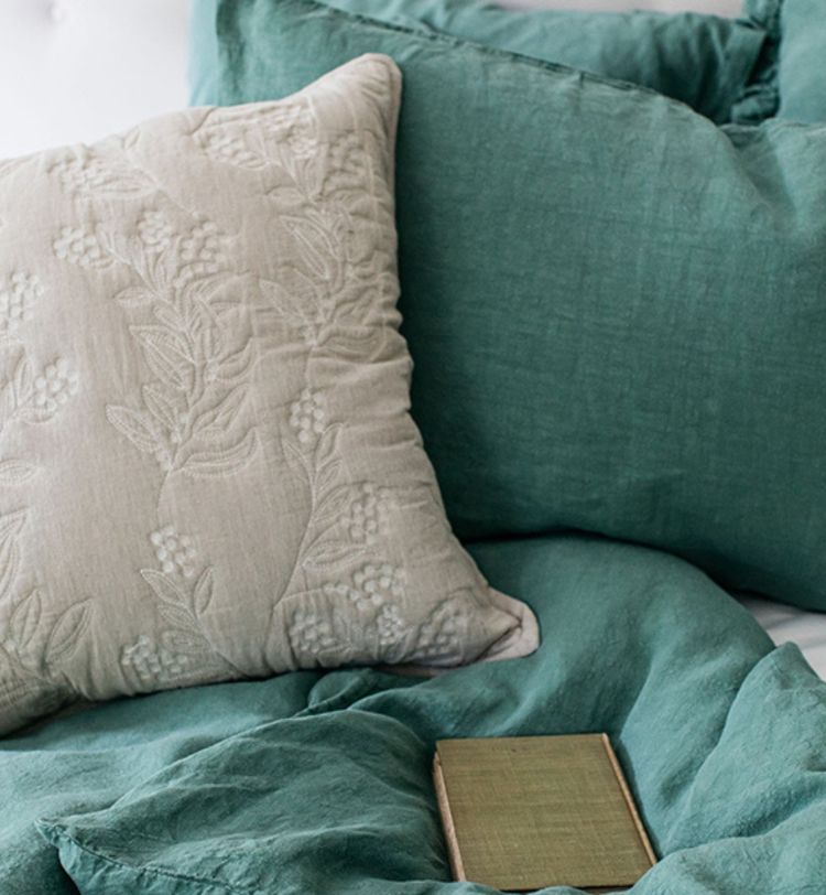 Green and beige pillows with floral embroidery on a bed with a matching green comforter. A closed book lies on the bed.