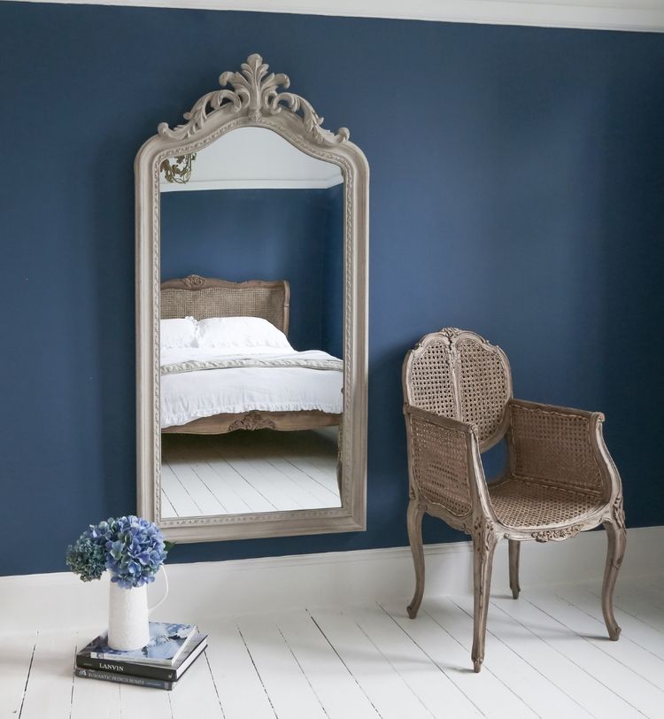 Elegant room with a large ornate mirror reflecting a bed, a wicker chair, and a small stack of books with a vase of blue flowers on the floor.