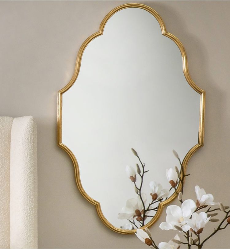 Ornate mirror with a gold frame on a beige wall, reflecting white magnolia flowers in the foreground.