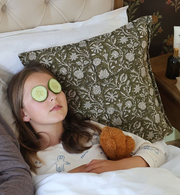 Girl relaxing in bed with cucumber slices on eyes, holding a teddy bear. Floral pillow and skincare products visible in the background.
