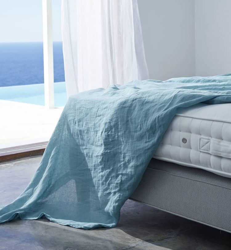 A light blue blanket drapes over a bed in a bright room with a sea view through a large window, sheer curtains partially open.