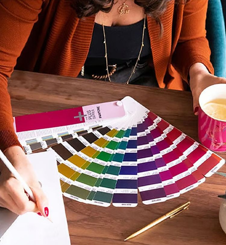 Person holding a pink mug and writing, with a colorful Pantone swatch fan spread out on a wooden table.