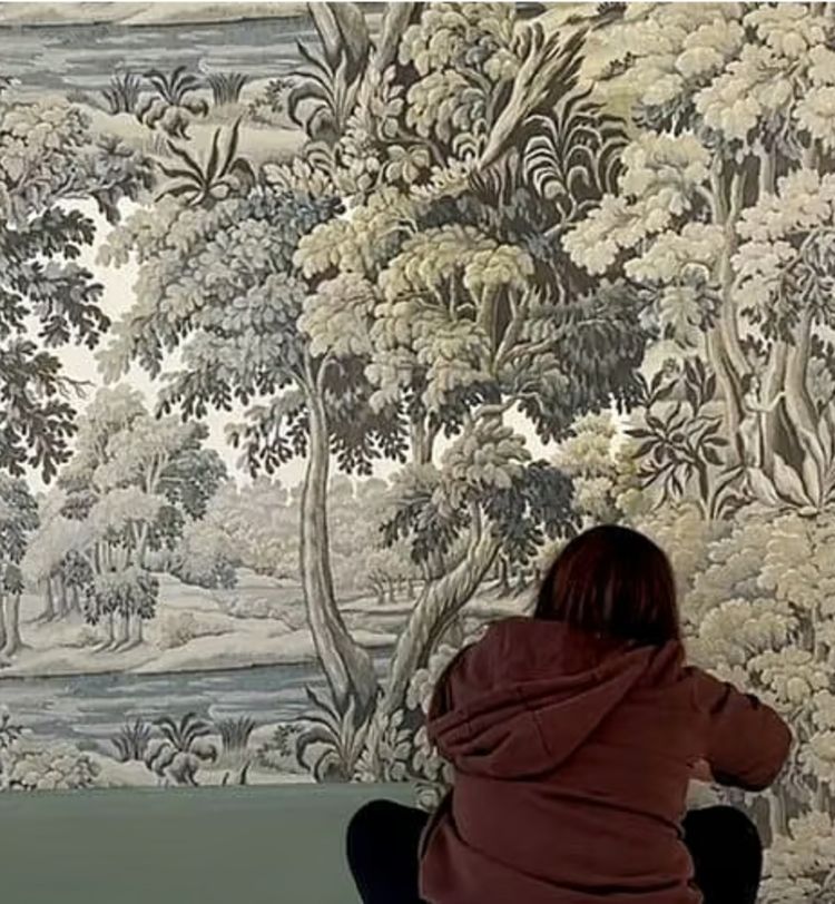 Person in a brown hoodie sitting and painting a large, detailed mural of a lush forest scene with trees and foliage.