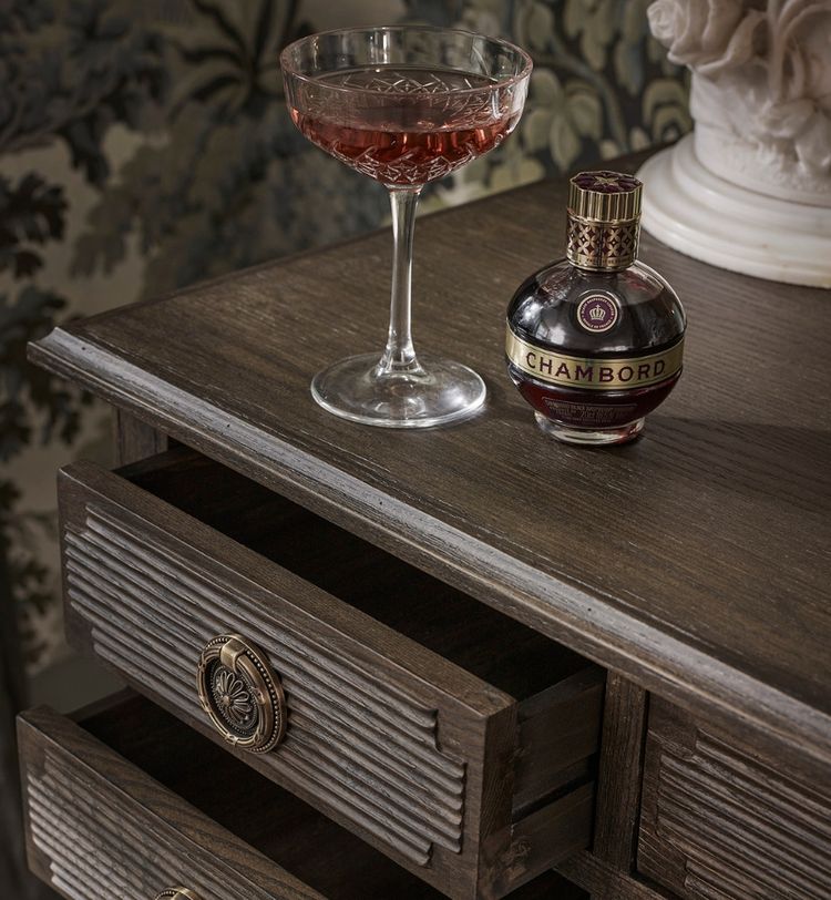 A glass of red liquid and a Chambord bottle sit on a vintage wooden dresser with open drawers, against a floral-patterned background.