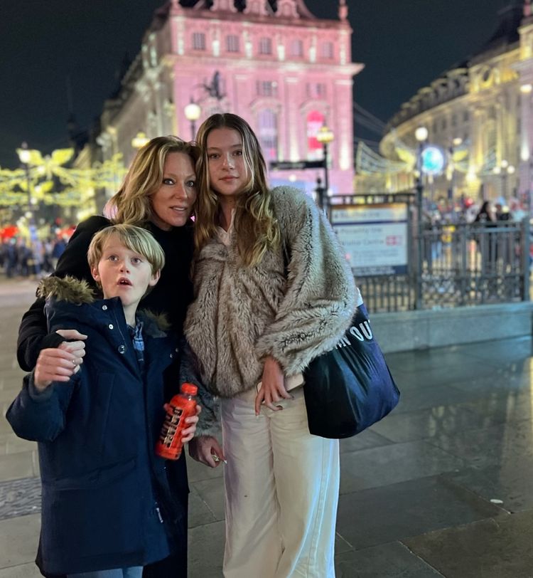 A woman and two children stand together in a brightly lit city square at night, with festive lights and a historic building in the background.