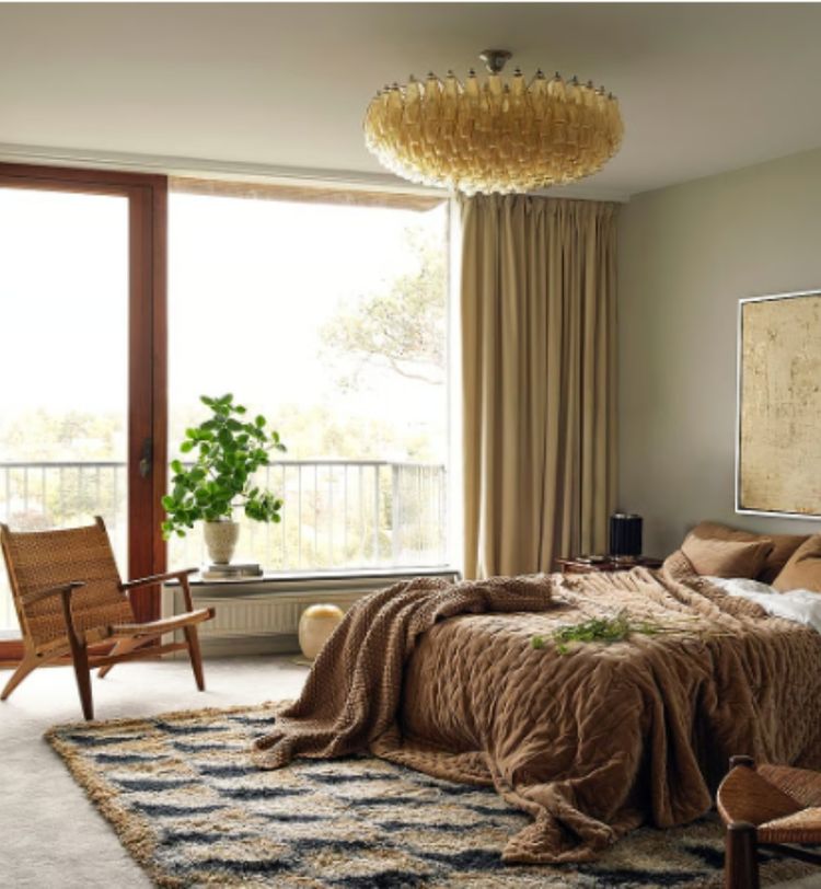 Cozy bedroom with a large window, beige bedding, a patterned rug, a wicker chair, and a leafy plant in a pot by the window.