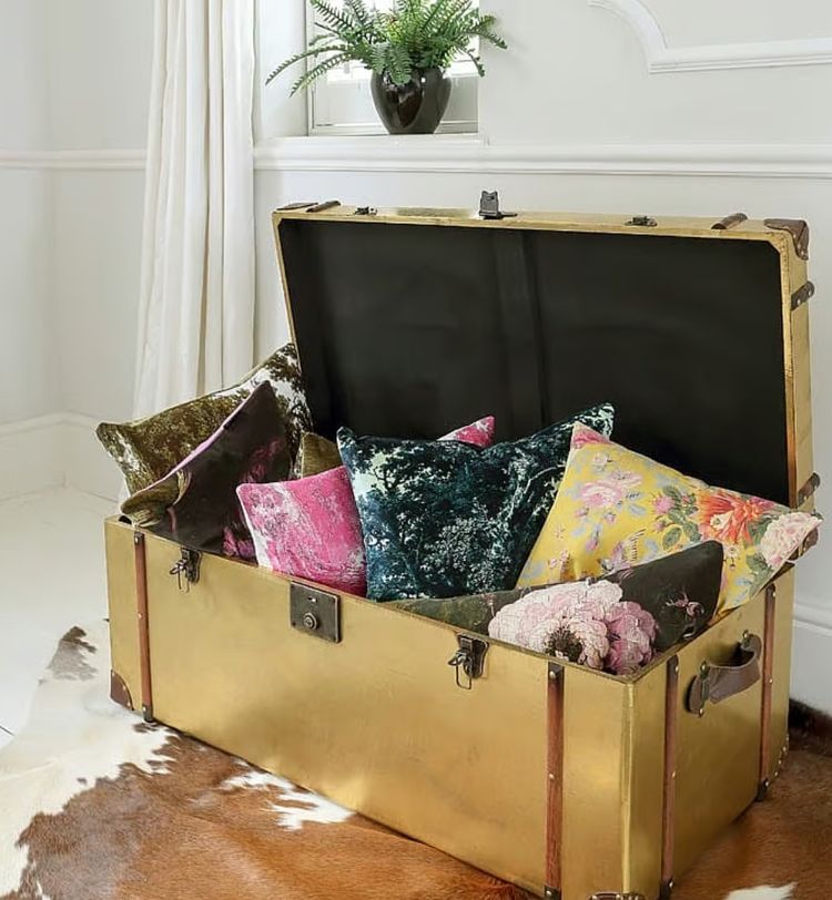 Open vintage trunk filled with colorful, patterned pillows. It rests on a cowhide rug in a bright room with a potted plant on top.