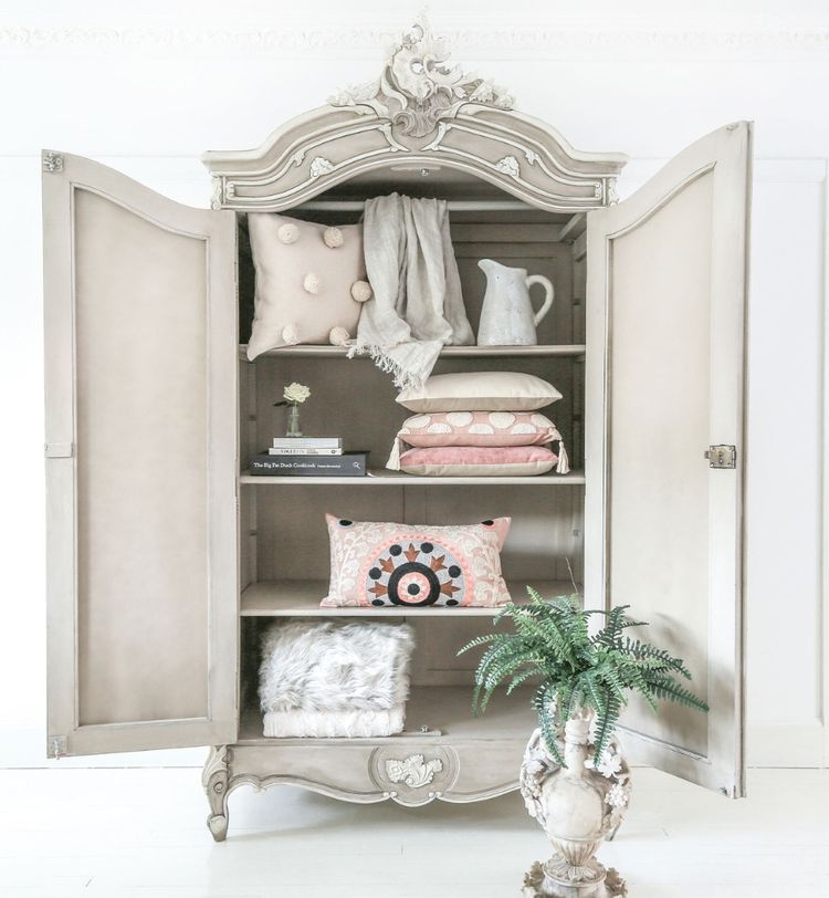 An ornate, vintage cabinet with open doors displaying neatly arranged pillows, a blanket, and a pitcher. A potted fern sits beside it.