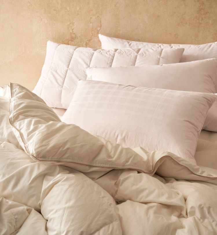 A cozy bed with soft, cream-colored pillows and a fluffy comforter against a beige wall, creating a warm and inviting atmosphere.
