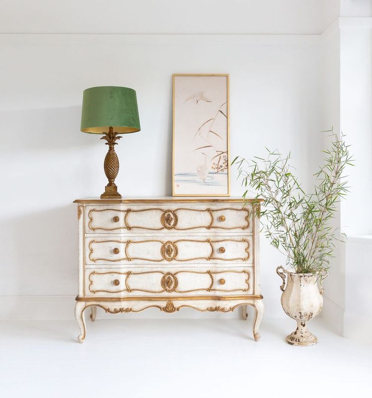 Vintage dresser with ornate details, green lamp, framed art, and potted plant in an elegant, minimalist room.