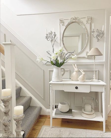 Elegant entryway with a white table, mirror, floral arrangement, and decorative bunnies. Candles adorn the staircase and table.