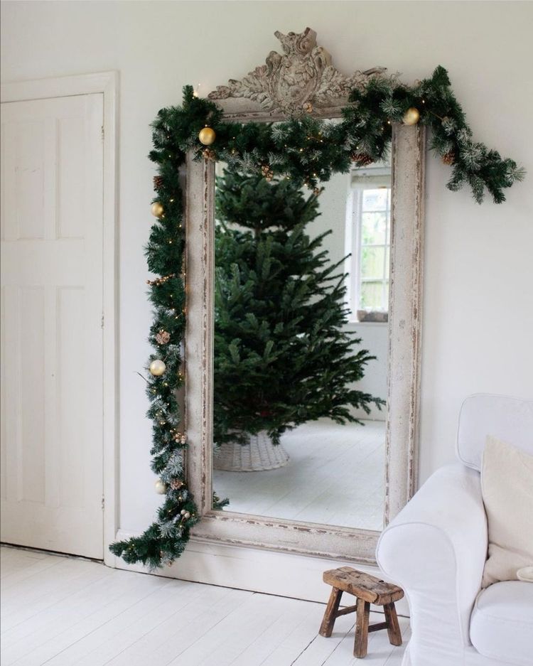 Ornate mirror with garland and gold ornaments, reflecting a Christmas tree. White room with a white sofa and a small wooden stool nearby.
