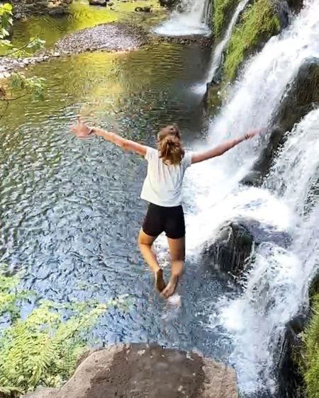 Person jumping into a clear river from a rocky ledge near a waterfall, surrounded by lush greenery.