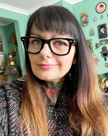 Woman with glasses and tattoos, wearing a speckled sweater, stands in a room with green walls adorned with various decorations.