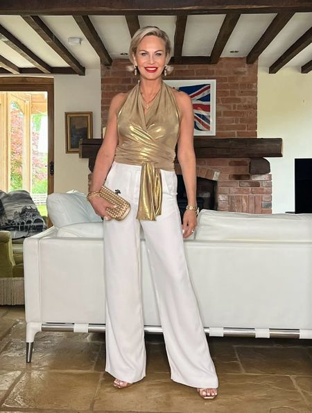 Woman in a gold halter top and white pants stands in a stylish living room with a brick fireplace and a Union Jack flag on the wall.