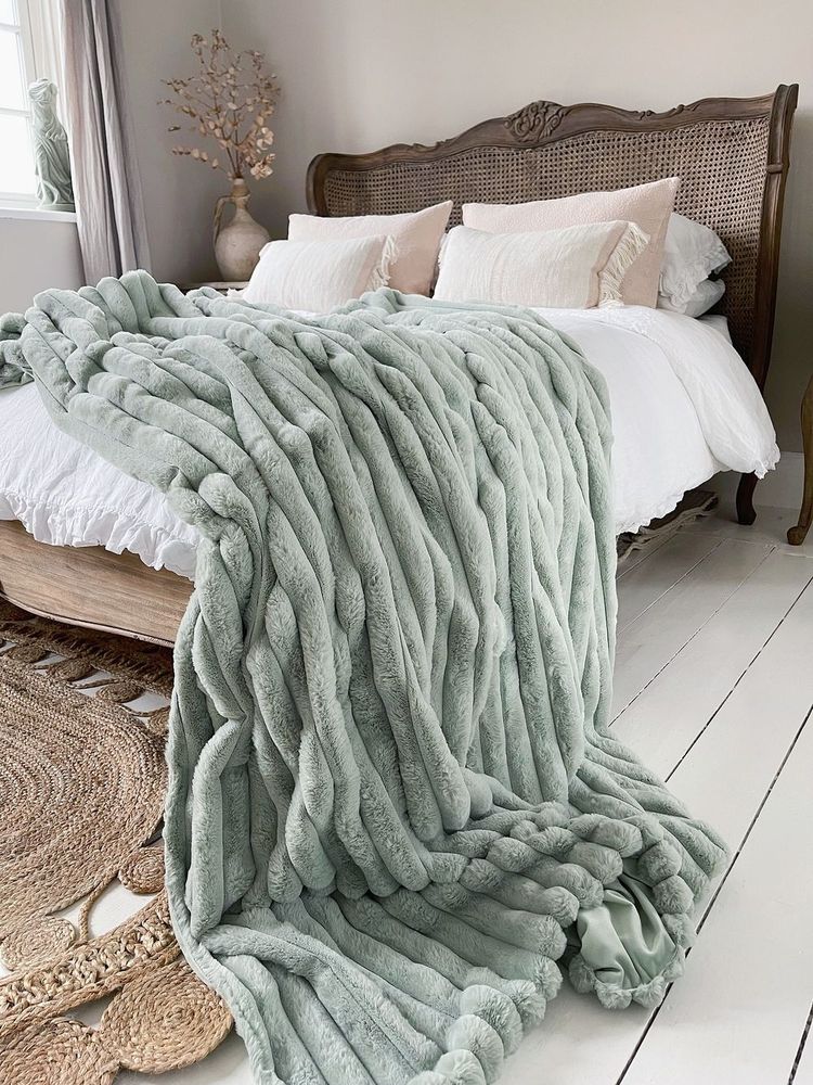 Cozy bedroom with a large, textured sage green blanket draped over a bed with white pillows. Wicker headboard and natural decor accents.
