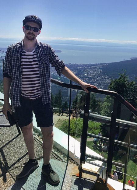 Man in sunglasses and casual clothes stands by a railing on a mountain, overlooking a cityscape and ocean under a clear blue sky.
