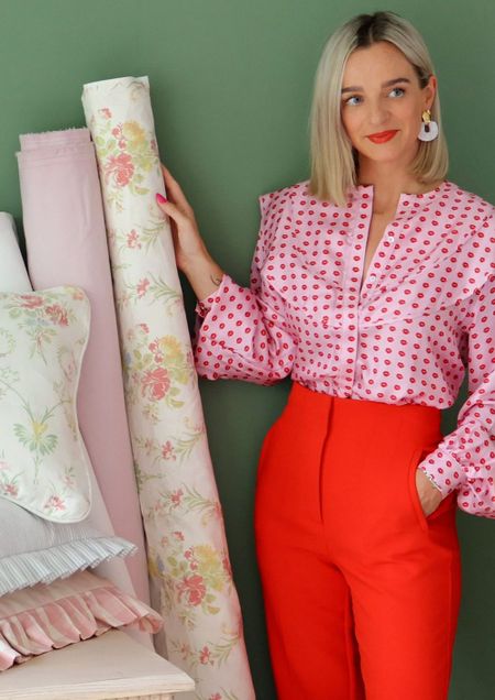 A woman in a pink polka dot blouse and red pants holds floral fabric rolls against a green wall, with stacked cushions nearby.