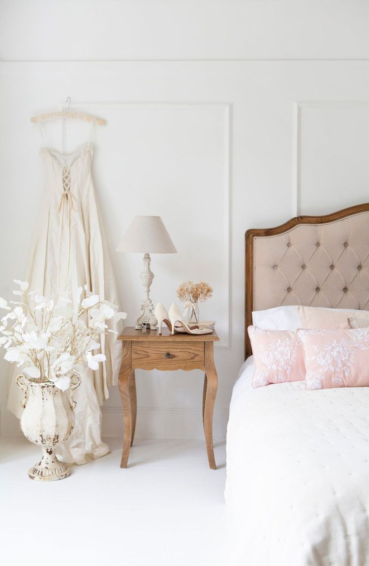 Elegant bedroom with a hanging wedding dress, wooden nightstand, lamp, pink pillows on the bed, and decorative flowers in a vase.