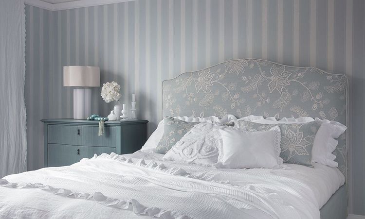 Elegant bedroom with a floral-patterned headboard, white bedding, teal dresser, and striped wallpaper. Decor includes a lamp and flowers.