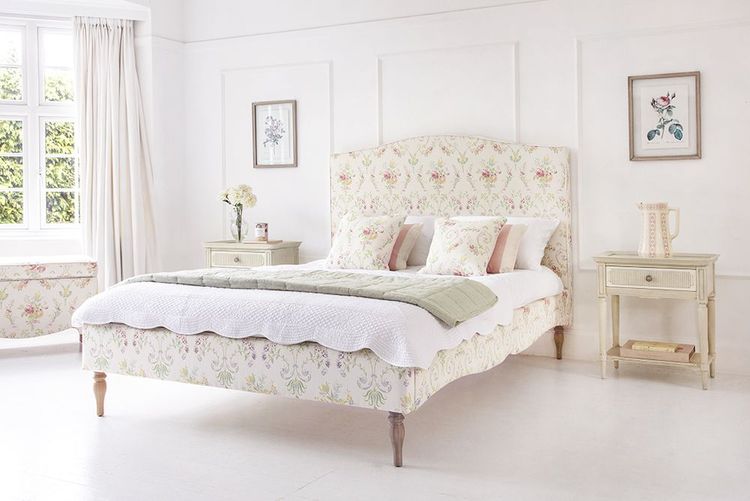 Elegant bedroom with a floral upholstered bed, white bedding, and pastel accents. Two framed floral prints adorn the light-colored walls.
