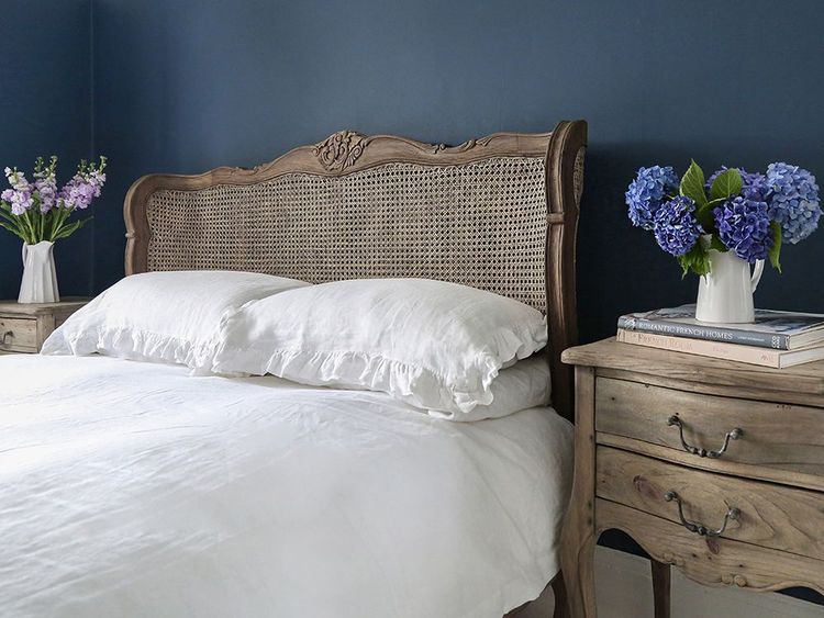 Cozy bedroom with a wooden bed, white bedding, and nightstands. Vases of purple and blue flowers adorn each side against a dark blue wall.