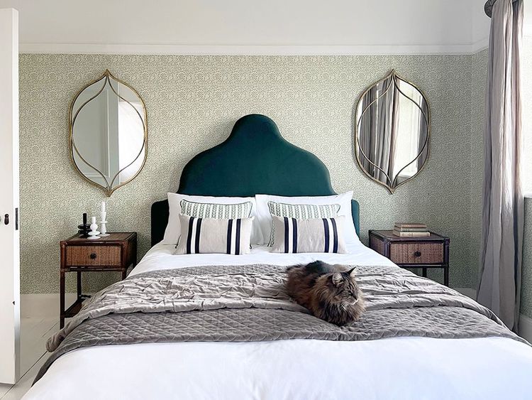 A cozy bedroom with a green headboard, two decorative mirrors, and a fluffy cat lying on a neatly made bed with gray bedding.