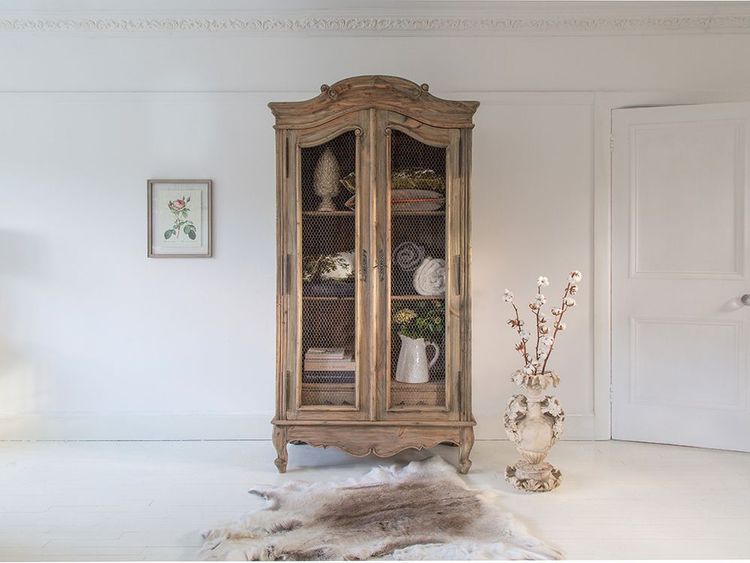 Vintage wooden cabinet with mesh doors, displaying decor items. Nearby, a vase with branches and a rug on the white floor. Framed art on wall.