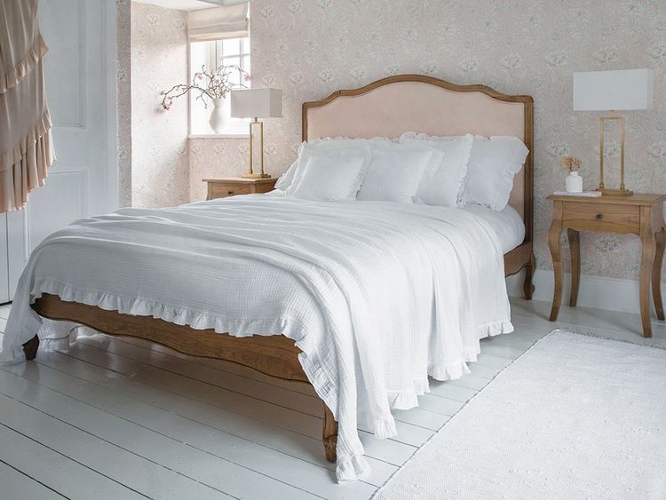 A cozy bedroom with a wooden bed frame, white bedding, floral wallpaper, and two wooden nightstands with lamps. Soft natural light fills the room.