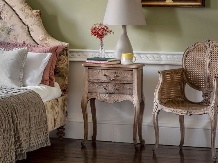 Cozy bedroom with a floral headboard, wooden nightstand, lamp, books, mug, and a wicker chair on a wooden floor.