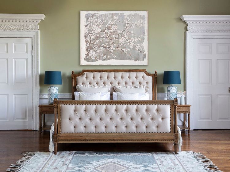 Elegant bedroom with a tufted bed, floral artwork, and matching side tables with blue lamps. White doors and a patterned rug complete the decor.