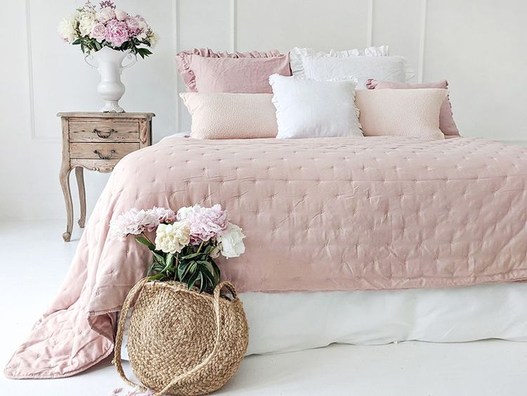 A cozy bedroom with a pink quilted bed, decorative pillows, a rustic wooden nightstand, and a basket of flowers on the floor.