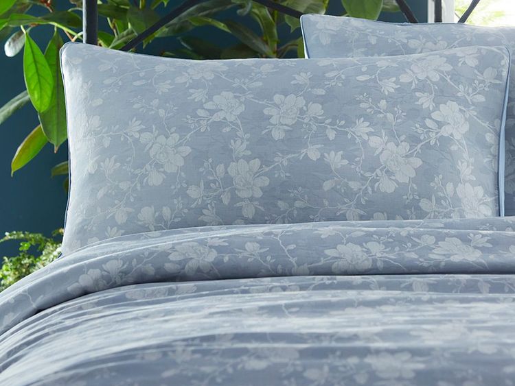 A bed with light blue floral-patterned pillows and comforter, set against a backdrop of green plants.