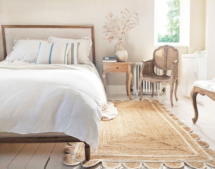 Bright, airy bedroom with a wooden bed, white bedding, rustic chair, side table, vase with branches, and a woven rug on light wooden floors.