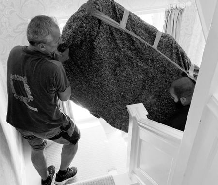 Two men carefully maneuver a large, wrapped sofa down a narrow staircase, showcasing teamwork and precision in a domestic setting.