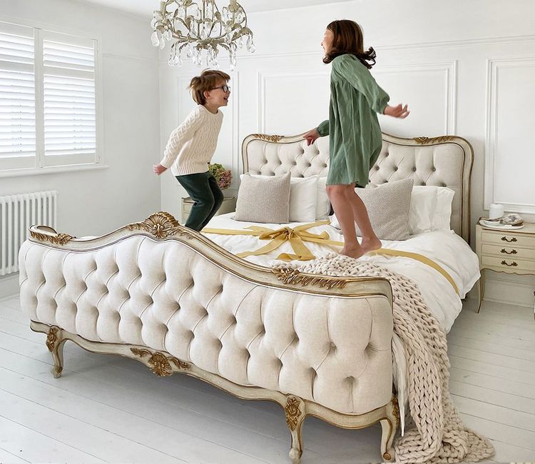 Two children joyfully jumping on an elegant, tufted bed in a bright, white bedroom with a chandelier and decorative pillows.