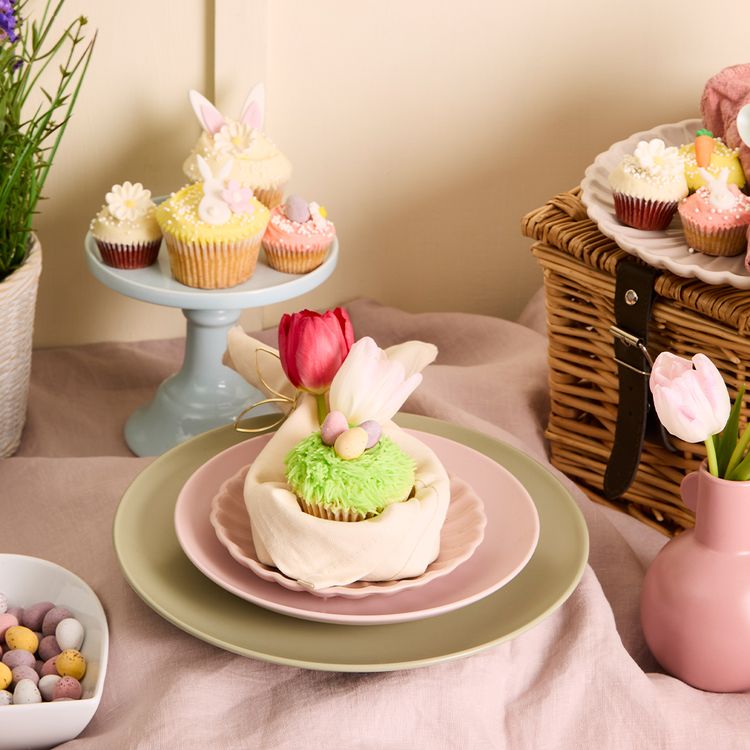 Spring-themed table setting with a cupcake, tulips, pastel eggs, and decorated cupcakes on a stand, basket, and flowers in the background.