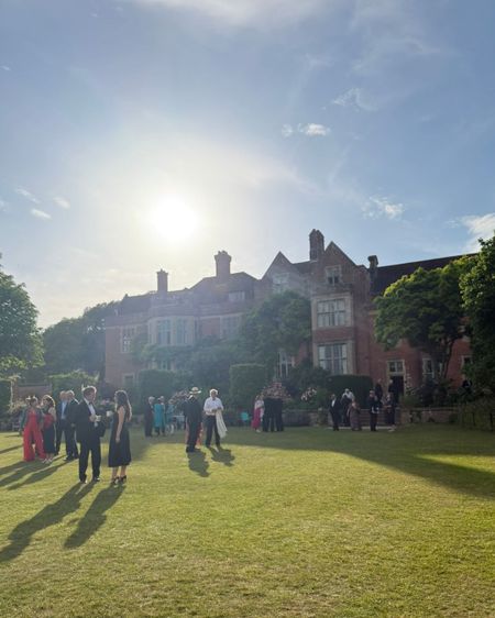 People in formal attire gather on a sunny day outside a large, historic brick building surrounded by greenery.