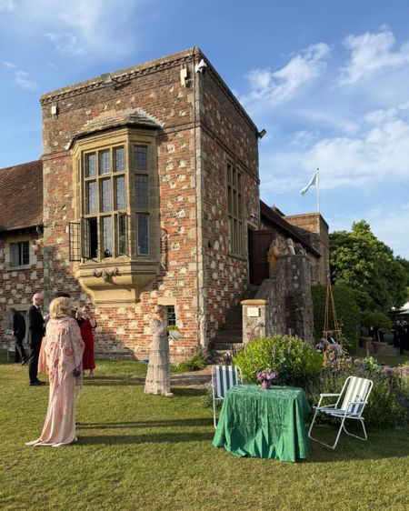 People in formal attire gathered outside a historic brick building at Glyndebourne, with a green table and chairs on the lawn under a clear blue sky.