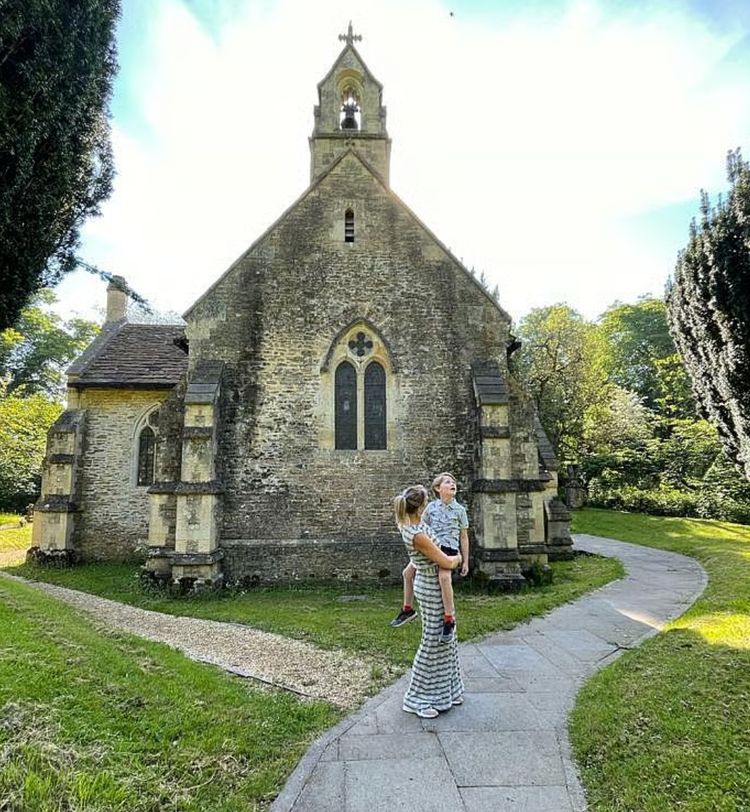 A person holding a child stands on a path in front of a small, old stone church surrounded by trees on a sunny day.