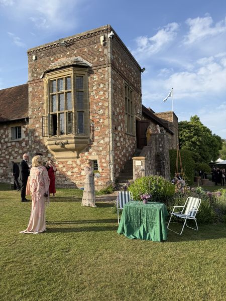 People in formal attire stand outside Glyndebourne's historic brick building. A green table with chairs is set on the grass. The sky is clear and sunny.