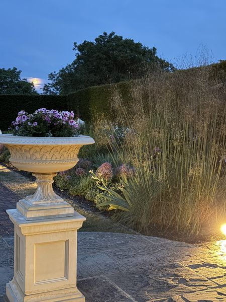 A garden scene at dusk with a stone planter of pink flowers, illuminated grasses, and a partially visible moon against a blue sky.