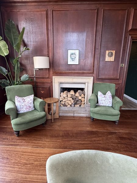 Cozy room with green armchairs, floral pillows, wooden walls, and a fireplace filled with logs. A potted plant and lamp stand nearby.