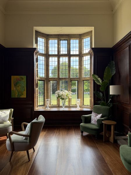 Cozy room with large bay window, wooden floor, green armchairs, and flowers on a wooden sill. Sunlight streams in, highlighting a serene ambiance.