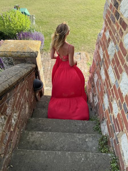 Georgia in a flowing red dress descends stone steps bordered by red brick walls, with green lawn and lavender plants in the background.