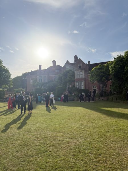People in formal attire gather on the Glyndebourne grounds under a bright sun and clear blue sky.