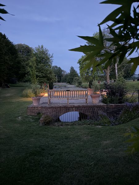 A wooden bench on a brick patio overlooking a garden, framed by trees and foliage under a blue evening sky.