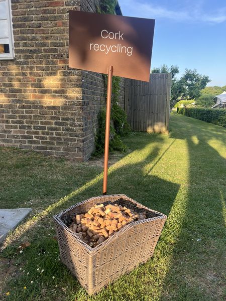 A wicker basket filled with corks under a sign reading "Cork recycling" outside, near a brick wall and garden.