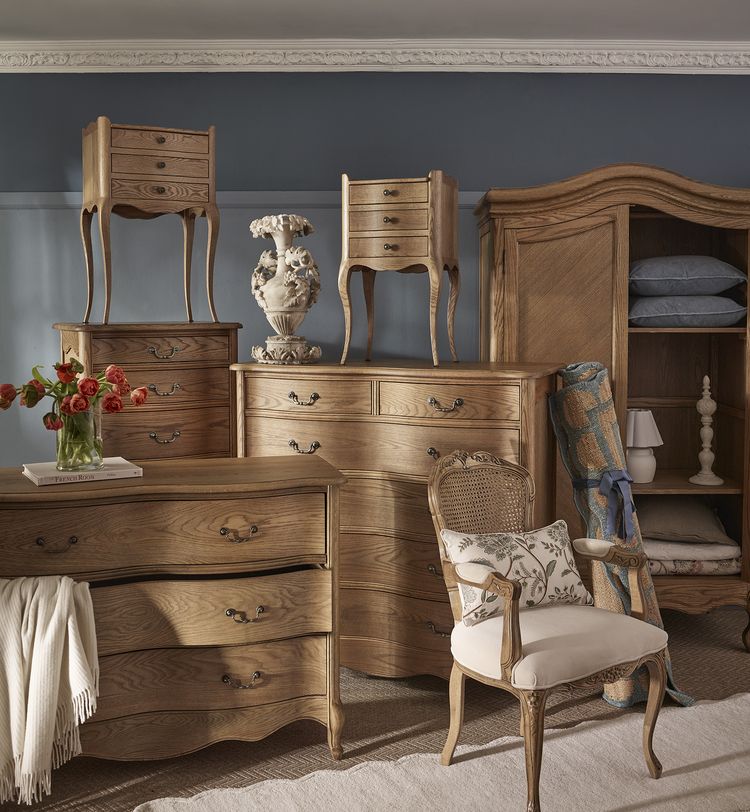 Elegant wooden furniture from the Love Story Collection set against a blue wall, featuring dressers, a chair, chests, bedsides, an armoire and multiple décor items.